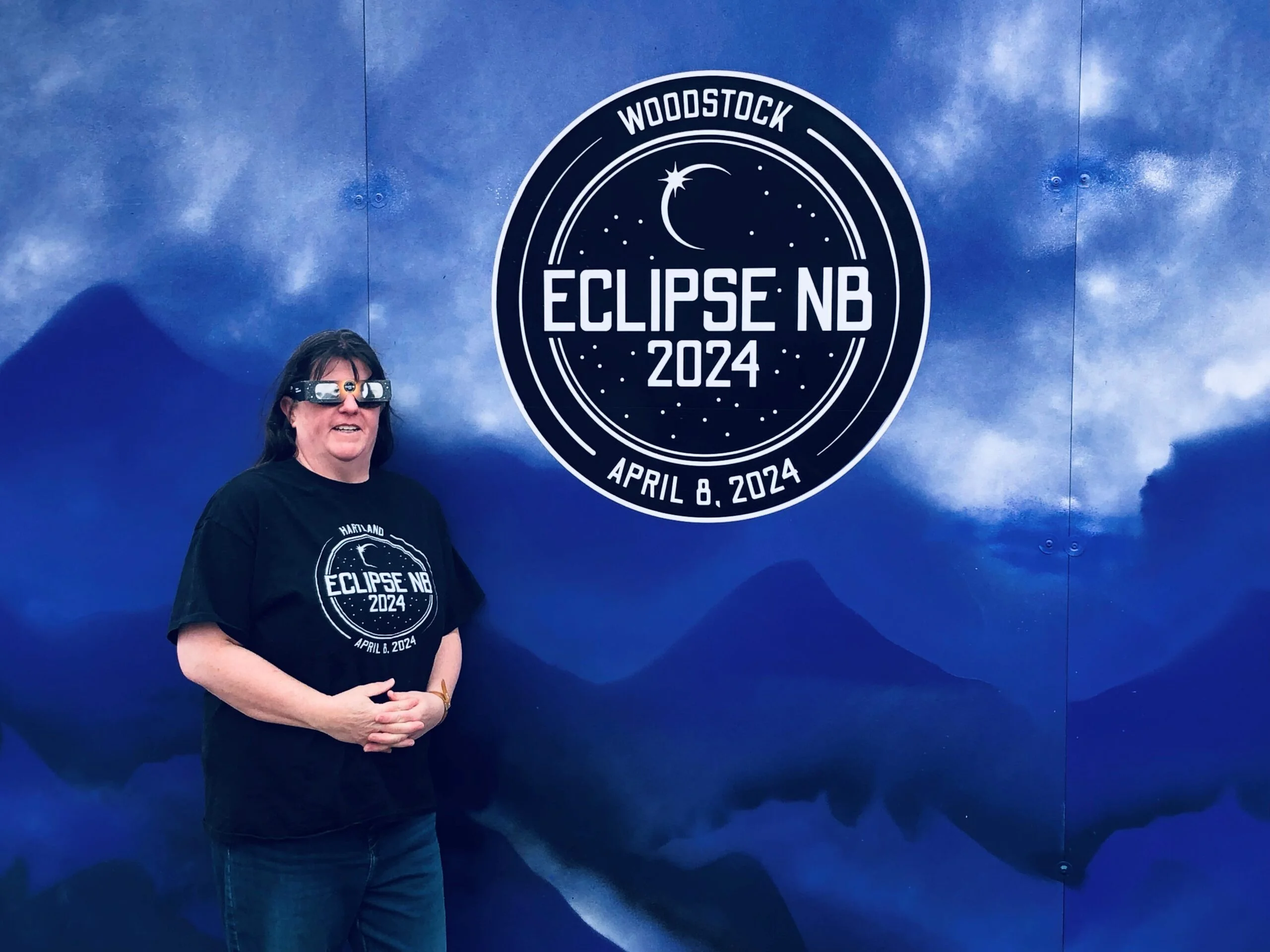 Photographer Tammey McLean, wearing her Eclipse t-shirt and approved eclipsed safety classes, stands next to the Eclipse mural in downtown Woodstock. (Jim Dumville photo)